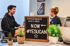 Student and health coach sitting behind a sign that says appointments available now #yesyoucan