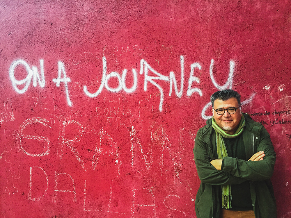 Santiago Vaquera-Vásquez standing in front of a red wall with graffiti that reads "on a journey"