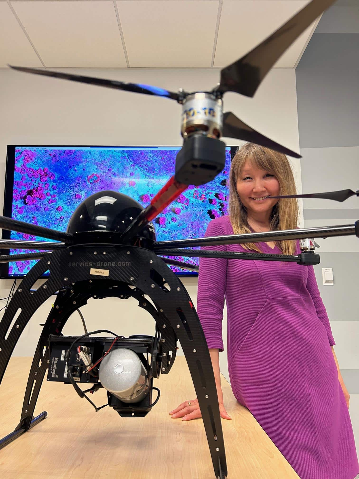Teki Sankey, with one of her lab's field drones in a classroom with a monitor behind her showing GIS mapping..