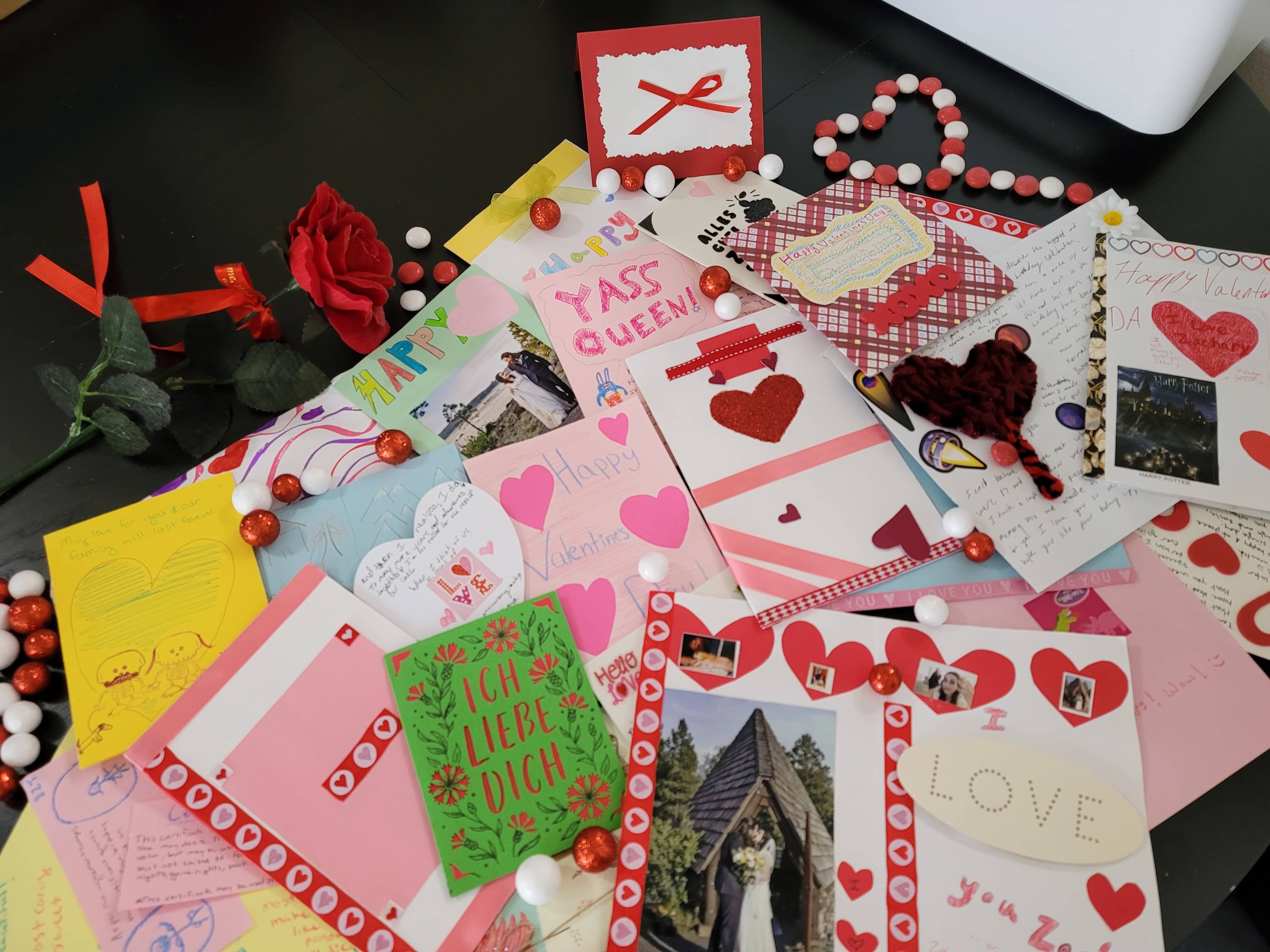 numerous homemade cards strewn about a table with a rose off to the side and red and white candies strewn about and in the shape of a heart.