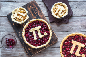 Two cherry pies with cherries visible and pastry with the Pi symbol sitting on top of cherries. Two smaller pies with choppped apples in them with the Pi symbol sitting on top. A small bowl of cherries off to the side.