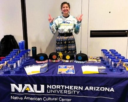 Rebekah McNiel at NAU’s Native American Cultural Center table with sway and informational sheets; McNiel doing the Lumberjack "L" with both hands.
