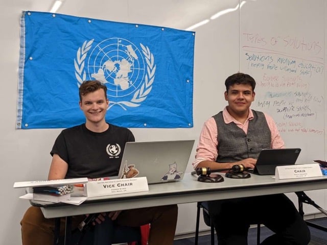 Two men sit at a table in front of a UN flag