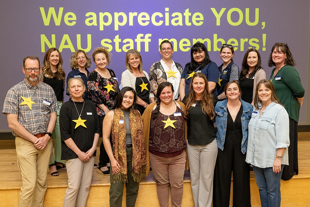Twenty people, including one man, stand in front of a sign that says "we appreciate you, NAU staff members!"