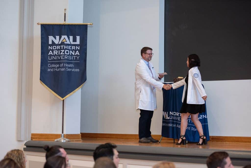 A woman in a white coat shakes the hand of a man in a white coat by a flag for NAU College of Health and Human Services