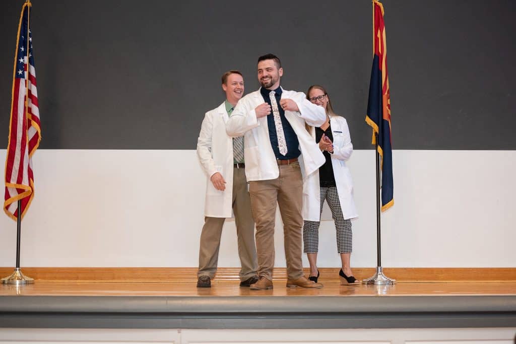 Two men and a woman wearing white coats smile on the stage.