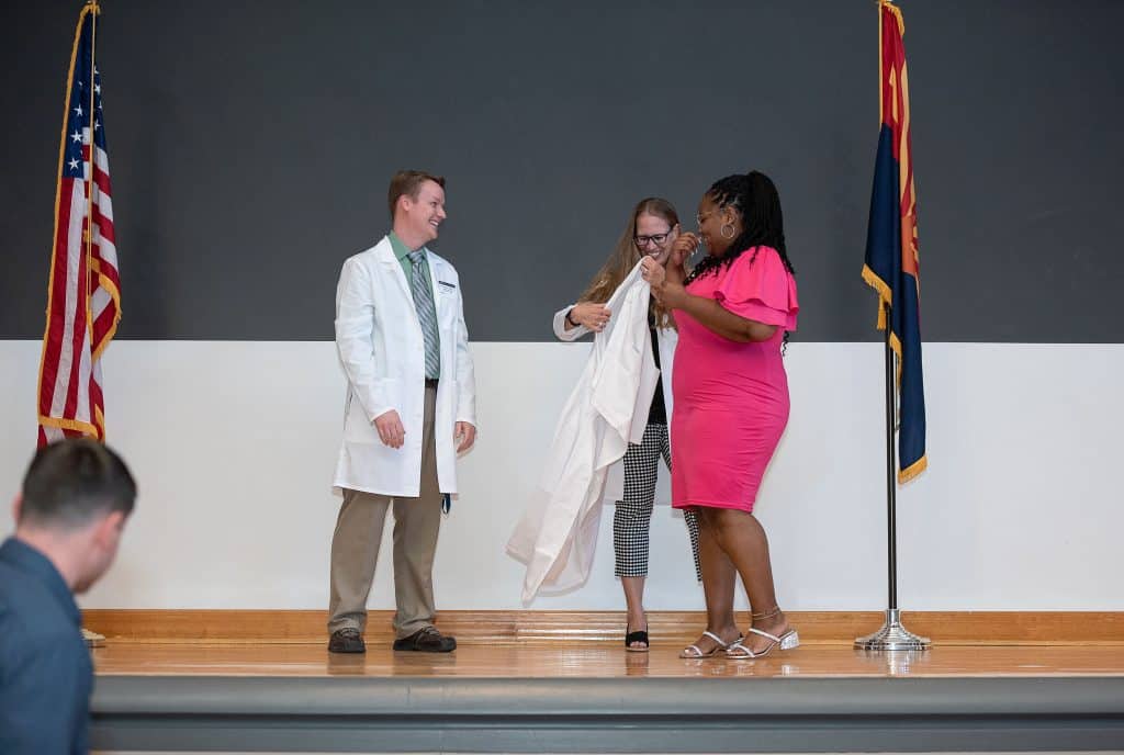 A man and a woman put a white coat on a woman in a pink dress.