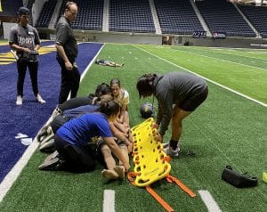 Athletic training students use appropriate techniques to put a manikin on a stretcher on a football field.