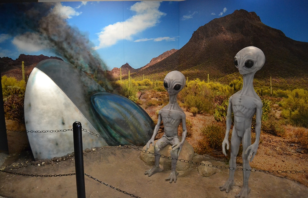 Two aliens by a crashed UFO in a museum exhibit.
