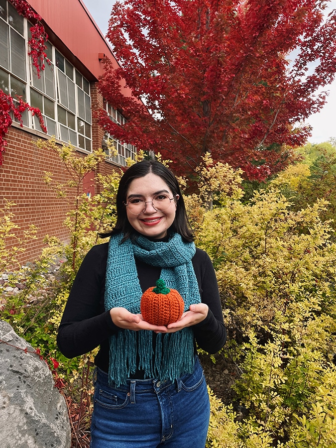 Kaylin McLiverty wearing a blue scarf and holding a knitted pumpkin in front of a tree with red leaves