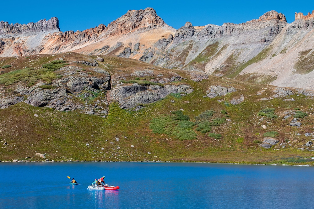 A small raft with people collecting data sits on top of a blue lake with mountains and sky in the background.