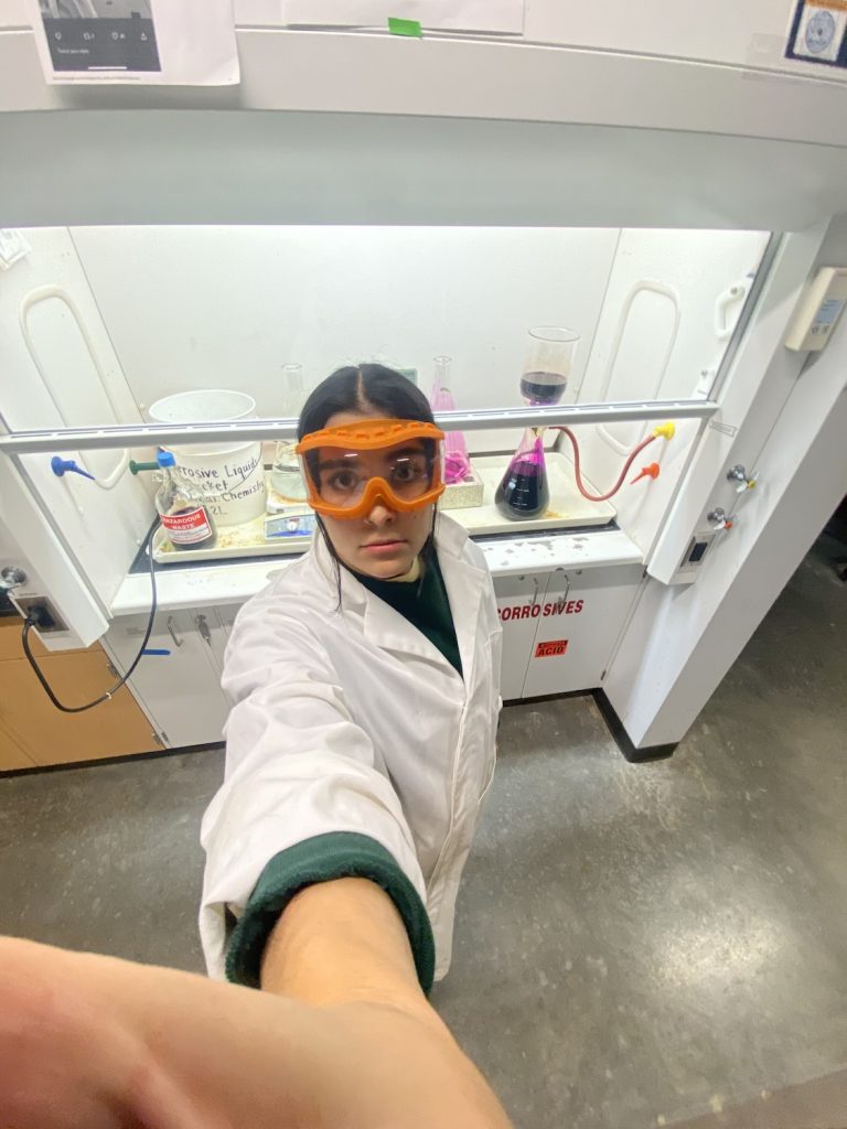 Selfie of Cecilia Perez wearing goggles and a white coat, with test tubes behind her