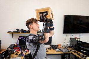 person wearing robotic arm attachments lifts a box in the air
