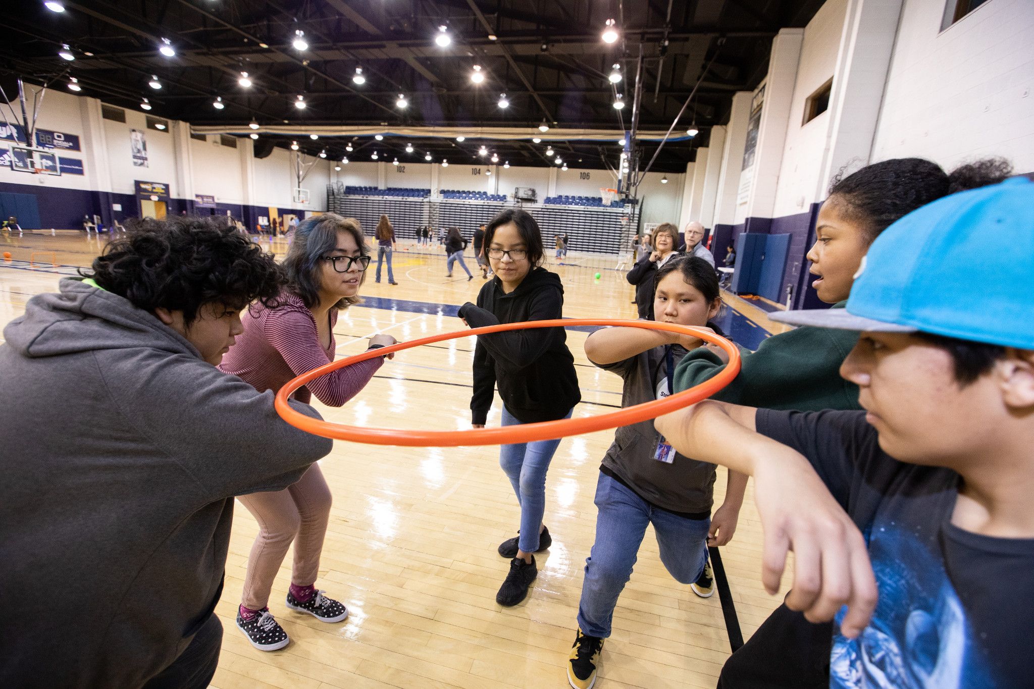 Young children standing in a circle with their hands on a hula hoop inside a gymnasium