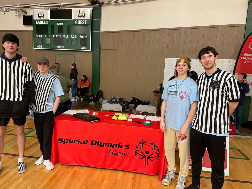 Four students dressed as referees posing at a Special Olympics Arizona table on a basketball court