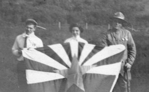 Historical photo of May Hicks holding up a flag with two men in uniform