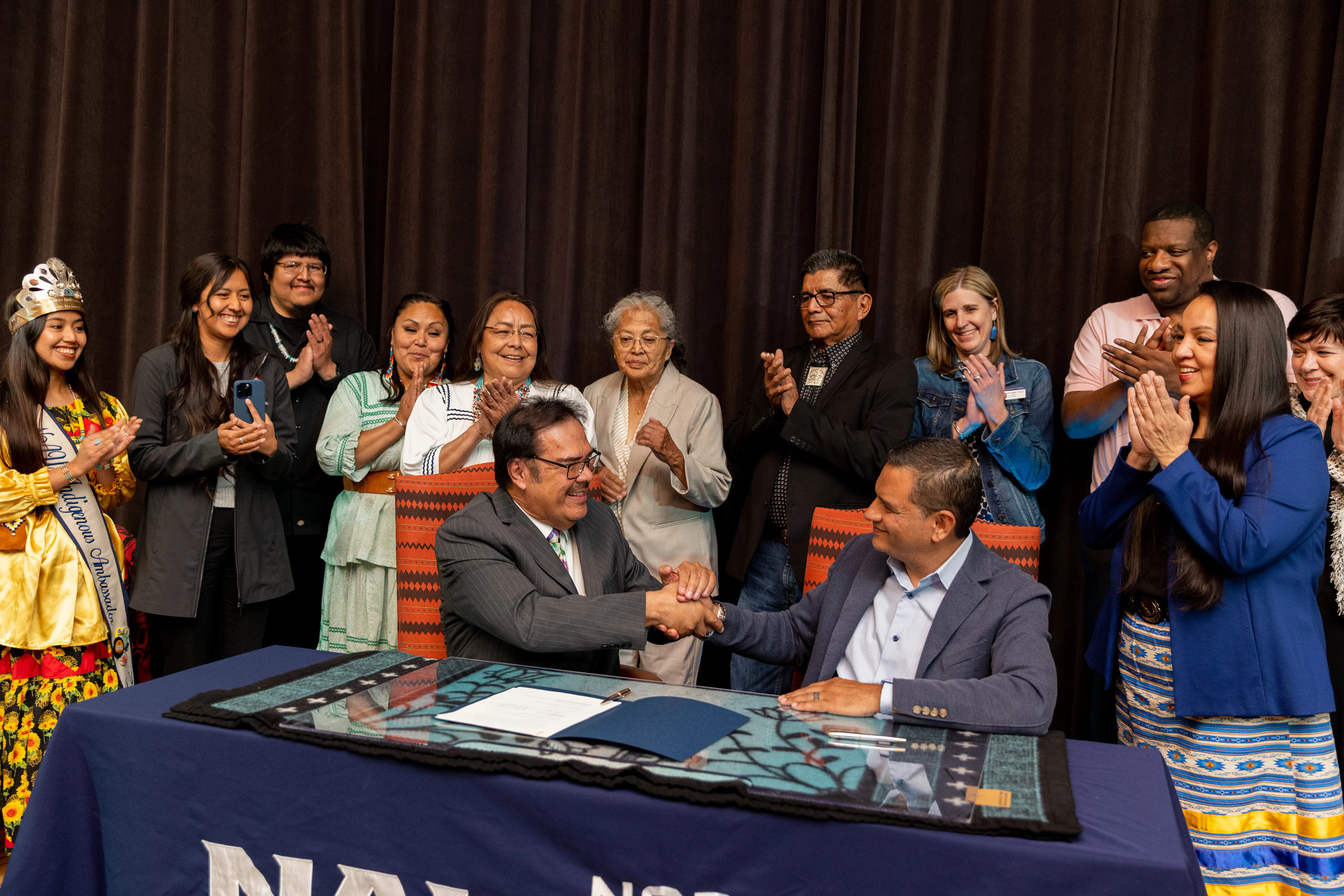 Martin Ahumada and José Luis Cruz Rivera shake hands while sitting at a table, while people in the background clap.