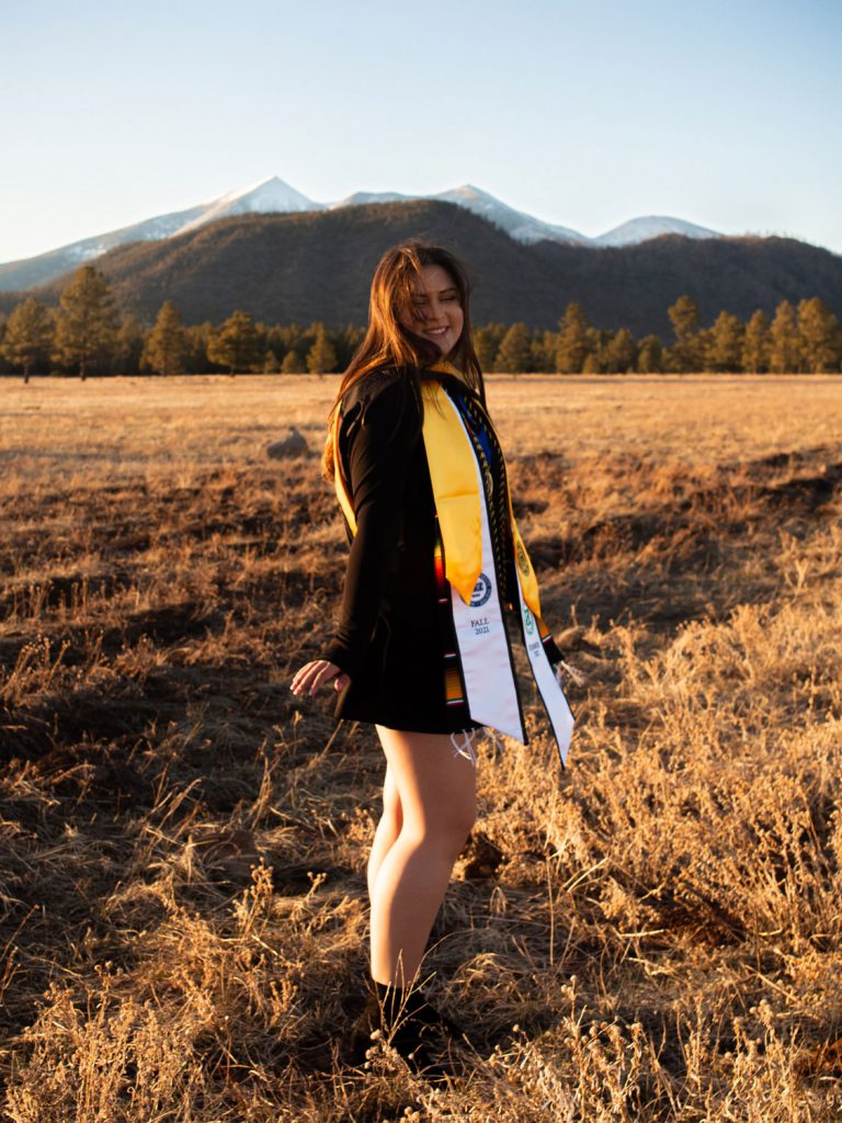 Hailey Lopez posing in front of the San Francisco Peaks in NAU graduation regalia