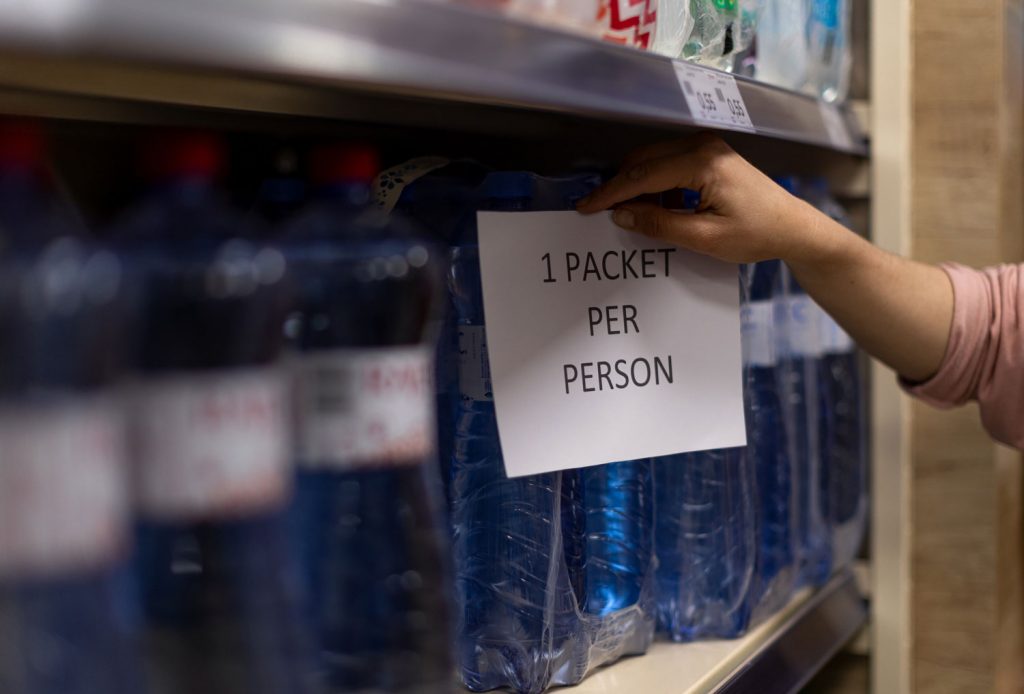 closeup of a shelf of water bottles with a woman reaching for a pack near a sign that says "1 packet per person"