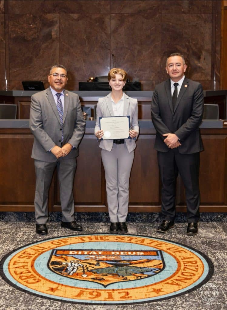 Goldmeer standing holding a certificate with two legislators