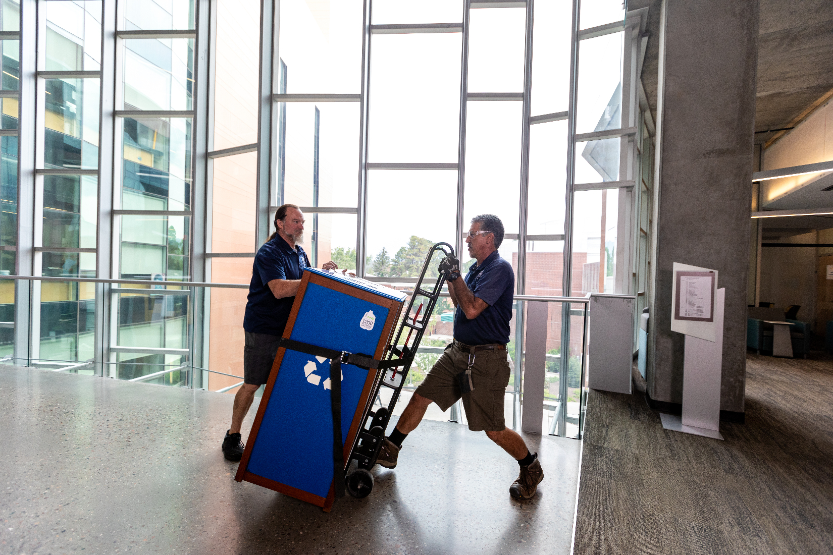 Two men use a dolly to transport a recycling bin.