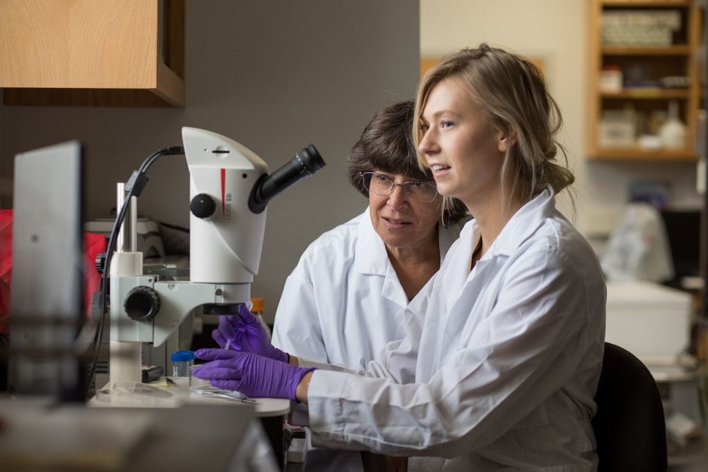 Catherine Propper looking over a student's shoulder while the student uses a microscope in a lab