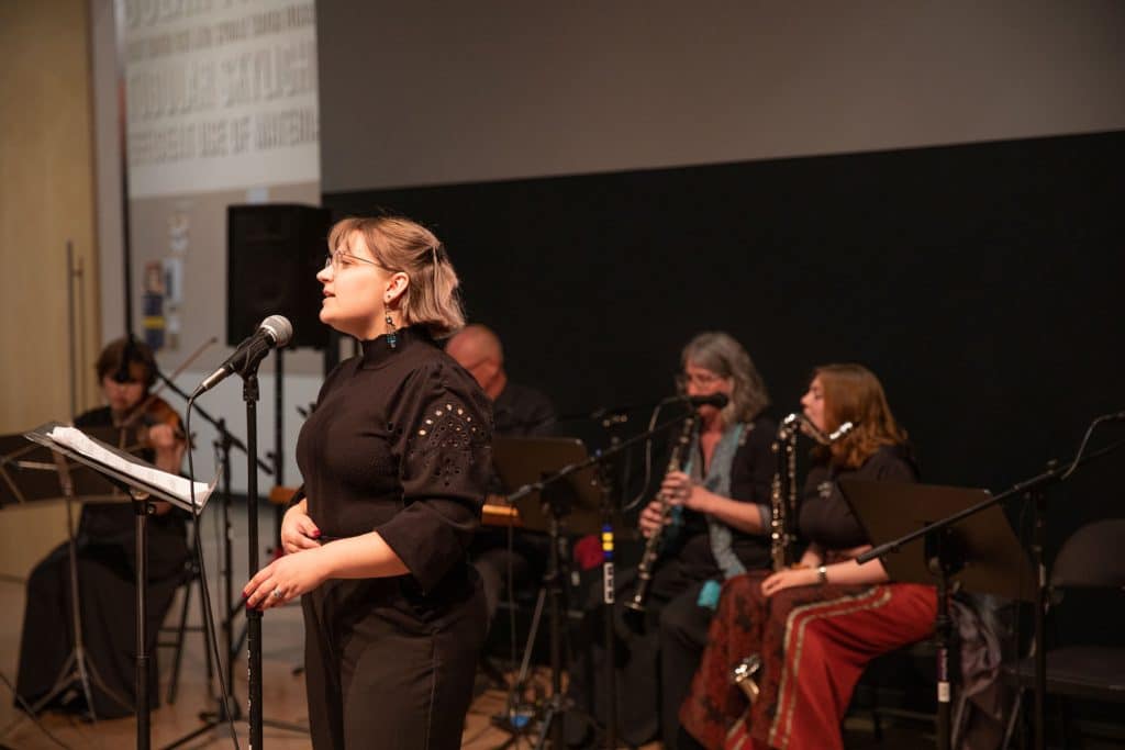 Woman singing at a microphone with two instrumentalists sitting behind her