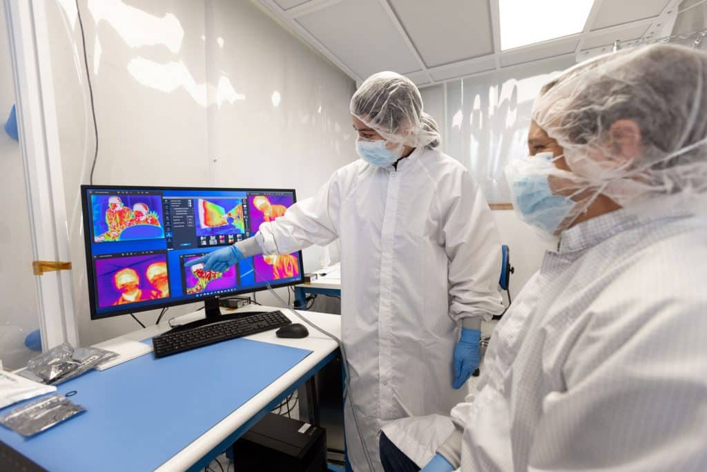 Two researchers in full-body PPE pointing to a monitor with infrared images