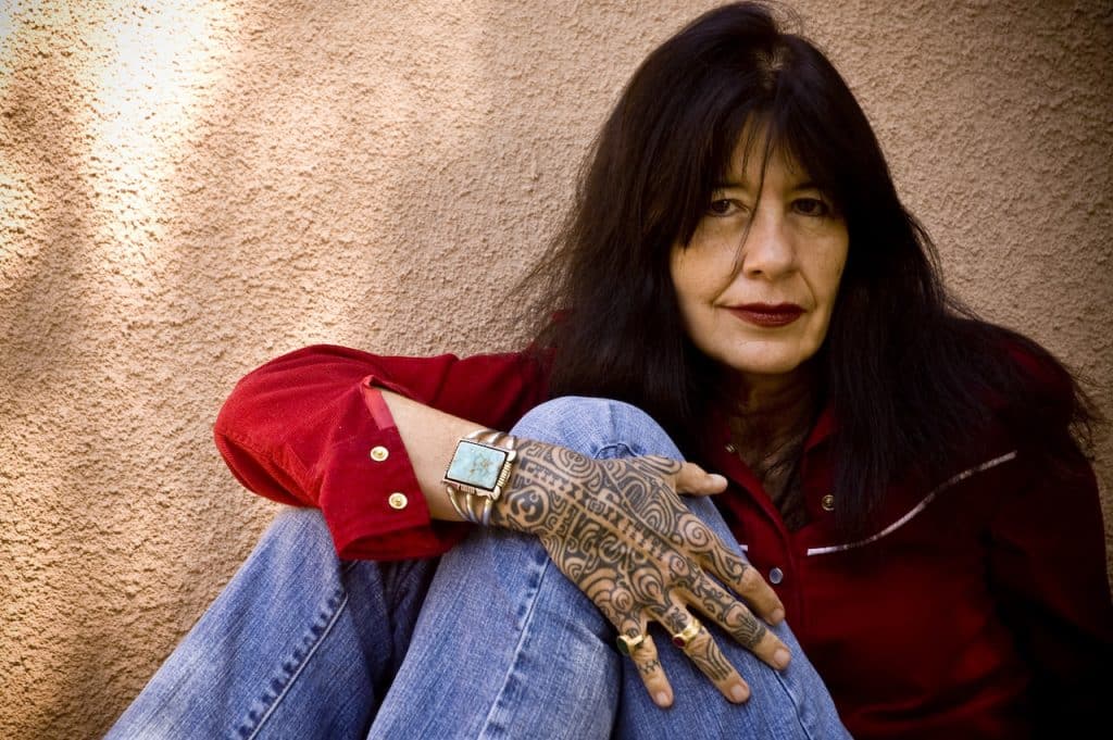 Headshot of Joy Harjo wearing a red shirt, jeans and a turquoise bracelet