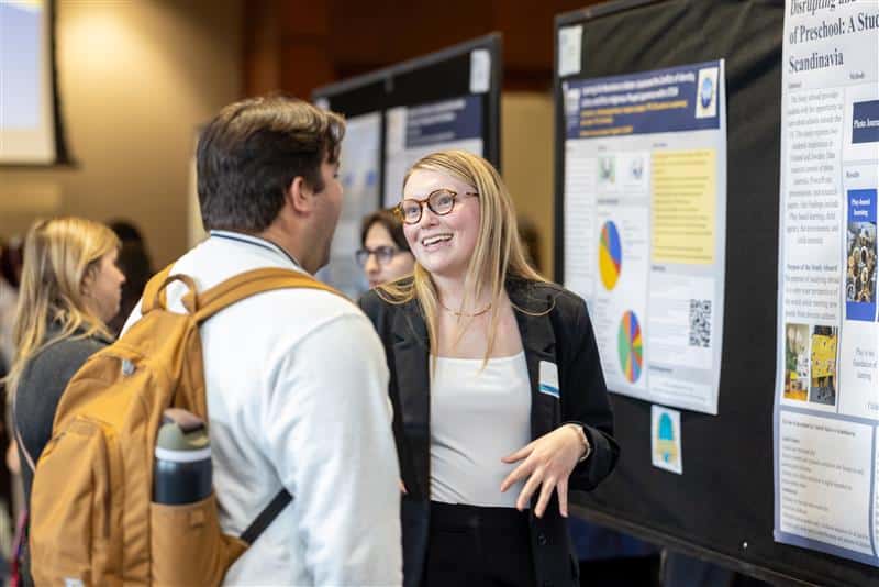 An undergraduate student speaks with a visitor in front of her symposium poster.