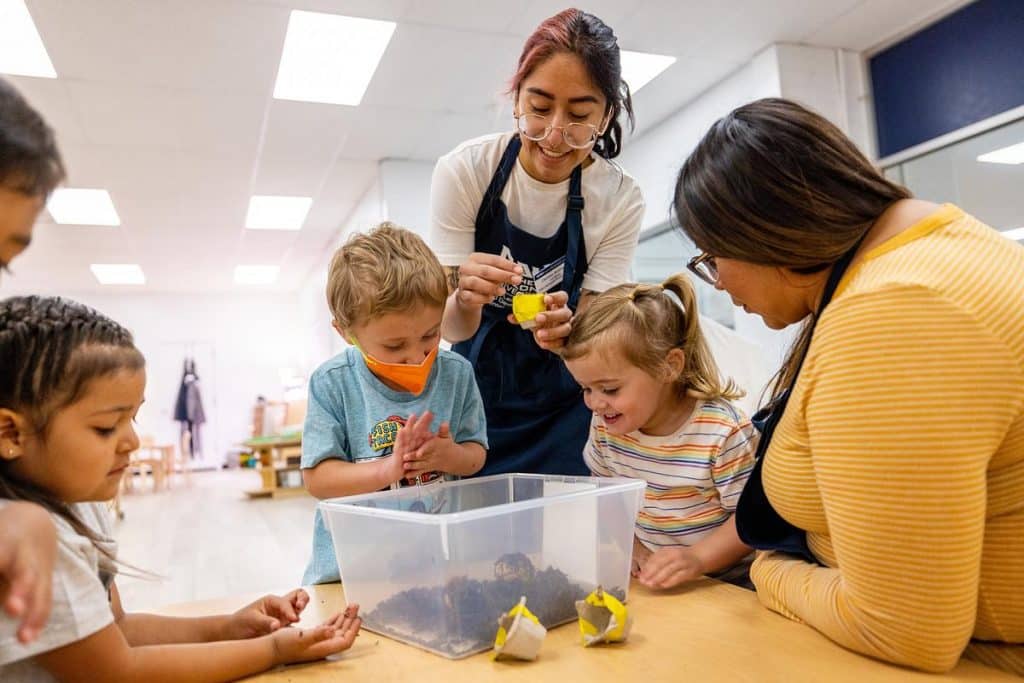 Teacher in an apron leaning over a table surrounded by kids