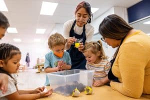 Teacher in an apron leaning over a table surrounded by kids