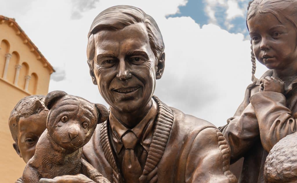 Bonze statue of Mister Rogers. He is holding a dog and kids are around him.