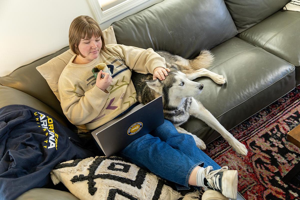 A woman does schoolwork from a laptop on her couch drinking coffee and petting a husky.