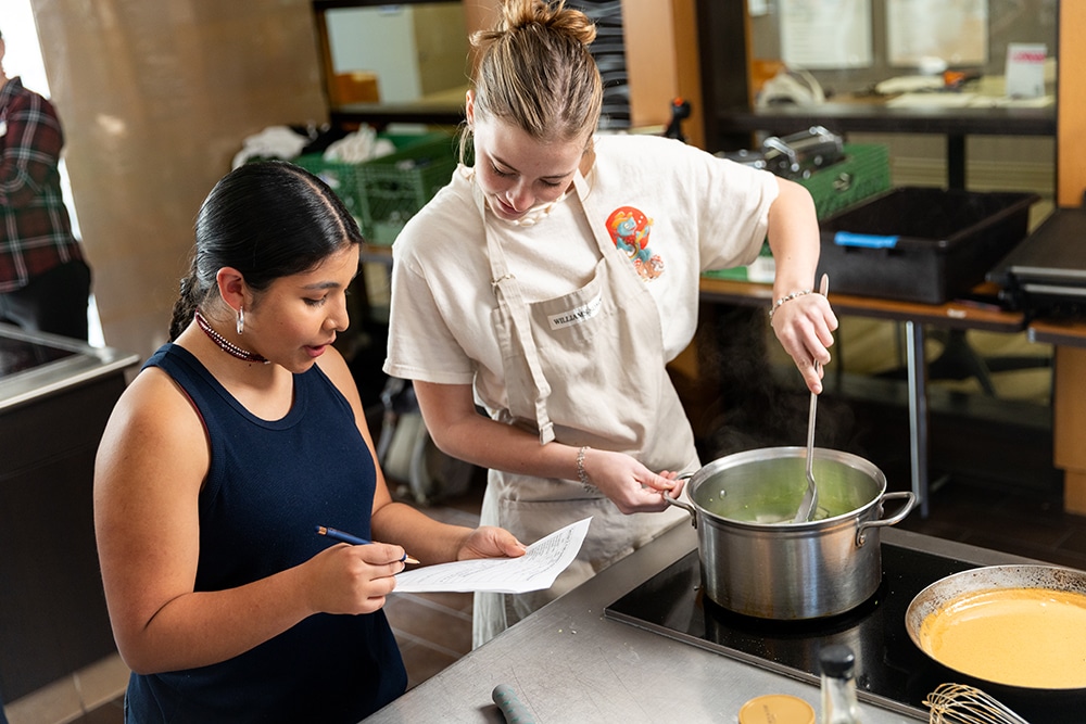 A student stirs food in a pot while another student asks questions.
