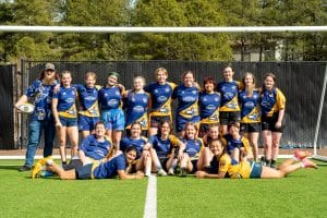The Lady Sharks rugby team members pose in their blue and gold uniforms on a rugby field.