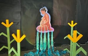 Art piece of pregnant woman who has water cascading down her body. The water goes down to water corn flowers around her.